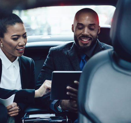 Multiracial adult colleagues in elegant clothes focusing on screen with interest and using digital tablet together while traveling in car